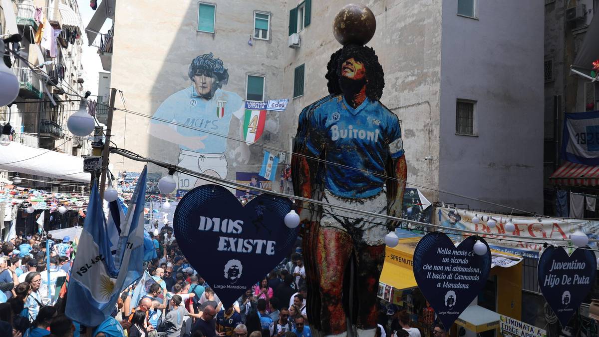 Pomnik Diego Maradony z piłką na głowie, w tle mural piłkarza i budynek.