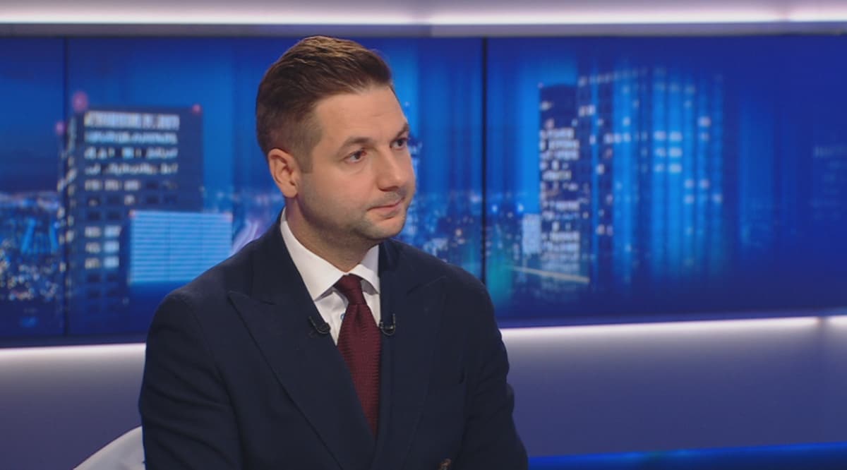 Mężczyzna w granatowym garniturze i białej koszuli z czerwonym krawatem siedzi, patrząc w bok. W tle znajduje się ekran z widokiem nocnego miasta.