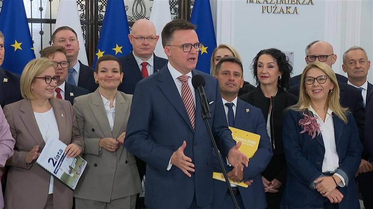 Grupa os&oacute;b, w tym mężczyzna przemawiający do mikrofonu, stojąca przed flagami.