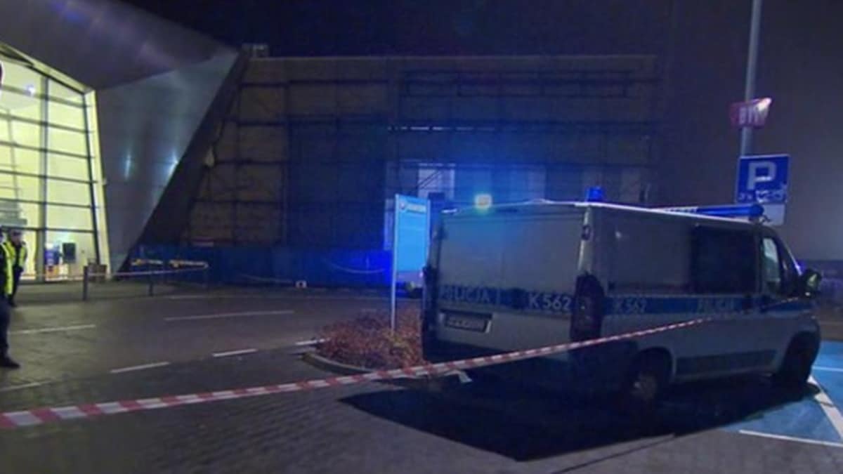 Centrum handlowe nocą z policyjnym furgonetką i taśmą odgradzającą teren.