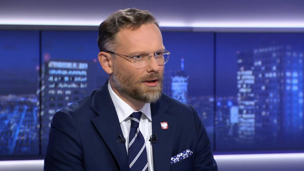 Mężczyzna w garniturze i okularach, siedzący na tle nocnej panoramy miasta.