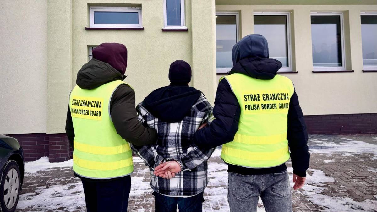 Trzech mężczyzn przed budynkiem Straży Granicznej, dw&oacute;ch w kamizelkach funkcjonariuszy, jeden zatrzymany.