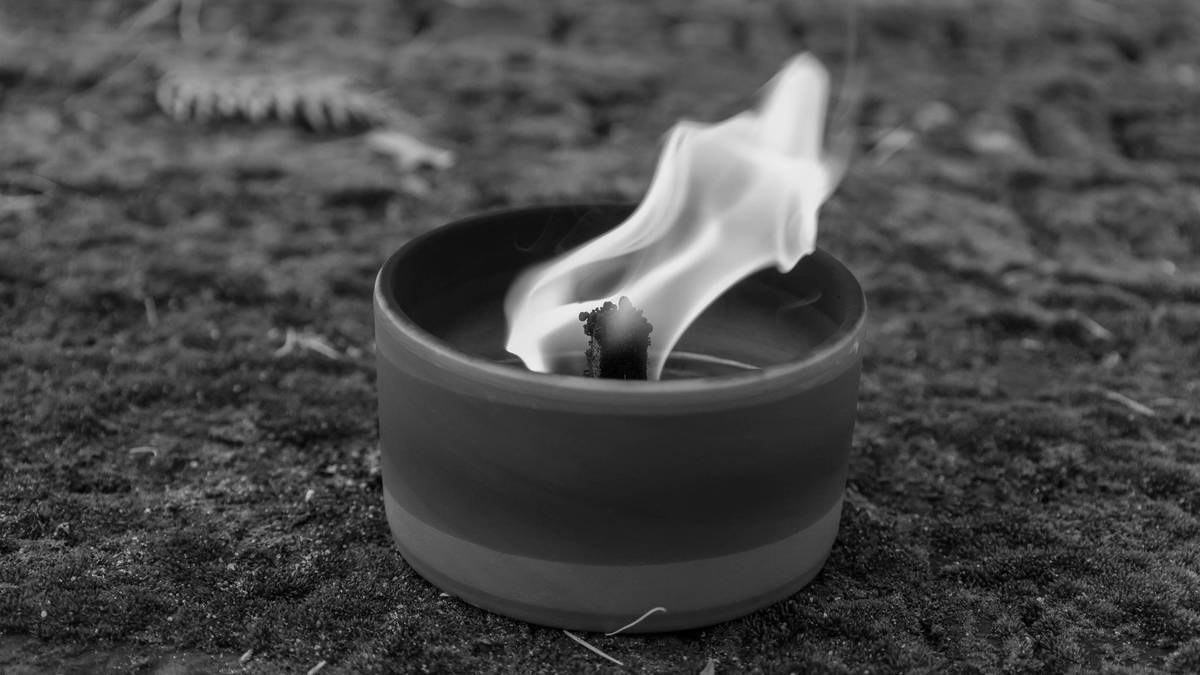 Płonąca świeca lub znicz w ceramicznym pojemniku na tle leśnej ści&oacute;łki.