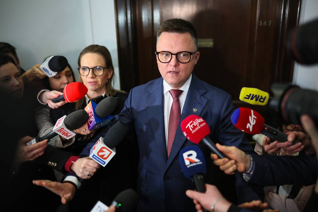 Szymon Hołownia otoczony przez dziennikarzy z mikrofonami podczas wypowiedzi w Sejmie.