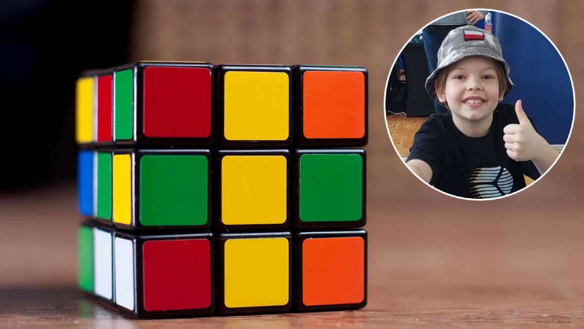 Częściowo ułożona kostka Rubika w żywych kolorach obok uśmiechniętego chłopca w kasku pokazującego kciuk w g&oacute;rę.