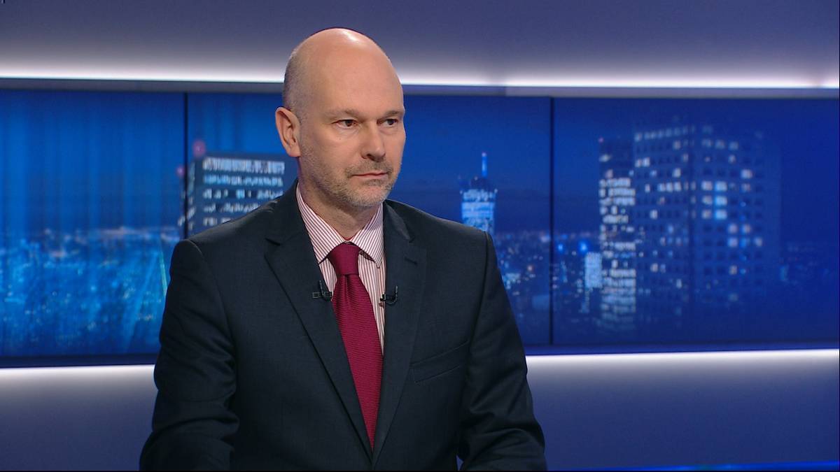 Mężczyzna w garniturze i krawacie siedzi na tle ekranów z panoramą miasta nocą.