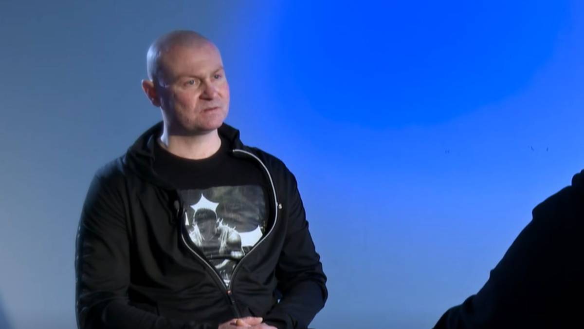 Mężczyzna siedzi, ubrany w czarną bluzę z kapturem na t-shirt z nadrukiem.