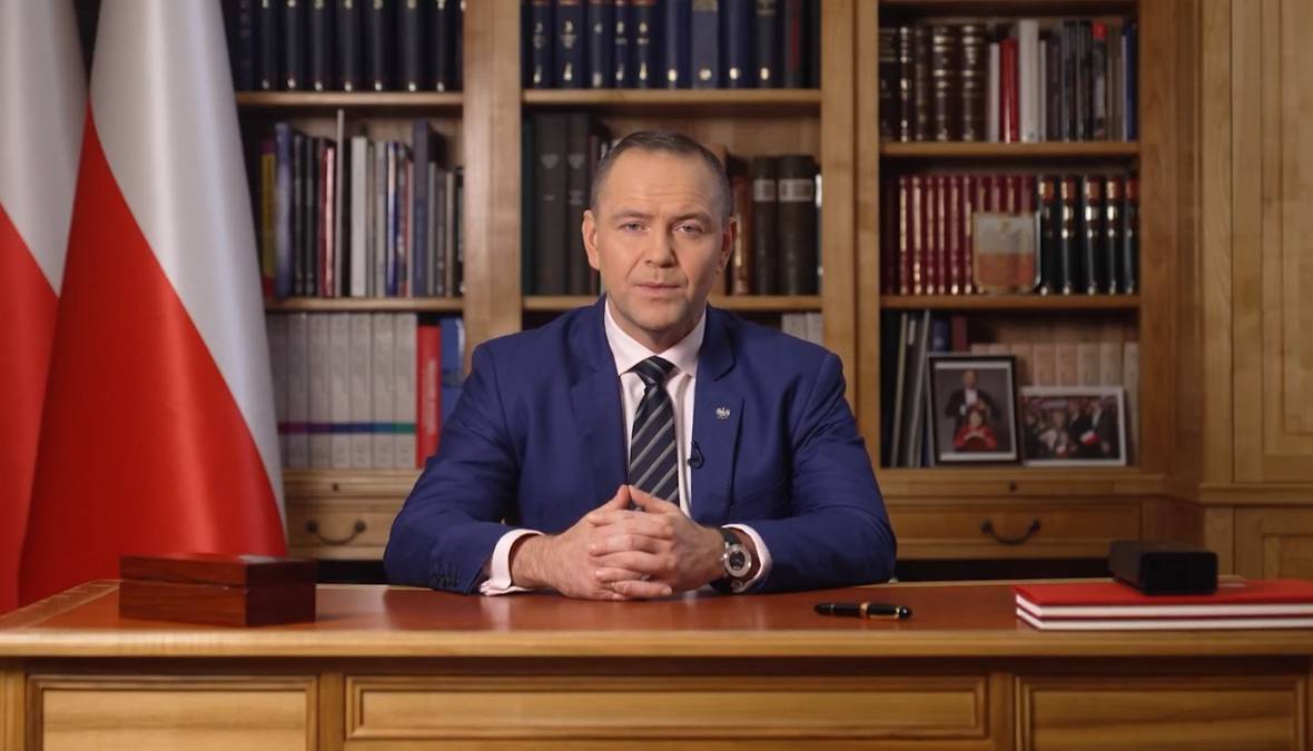 Prezydent Karol Nawrocki siedzi za biurkiem, przemawia w kierunku kamery. W tle znajdują się dwie polskie flagi i regały wypełnione książkami.