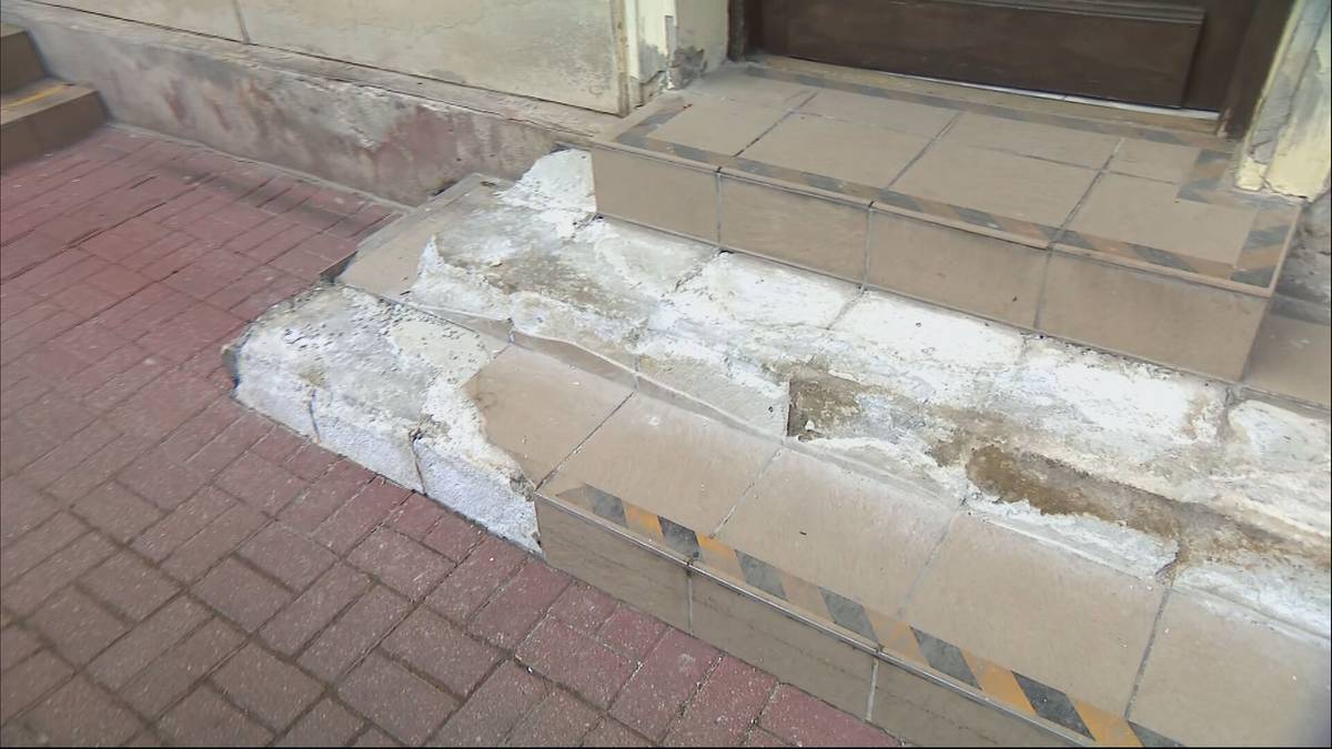 Trzy stopnie prowadzące do wejścia budynku, z widocznym uszkodzeniem tynku i płytek, obok fragment chodnika z czerwonej kostki.