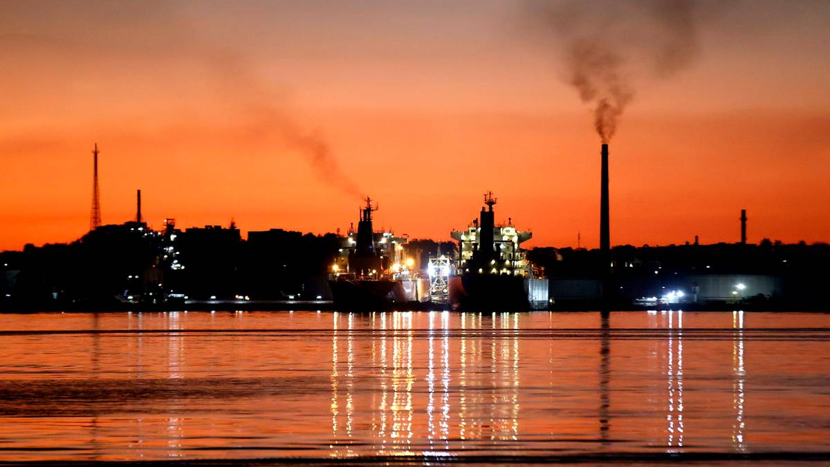 Sylwetka miasta z fabrykami i statkami na tle pomarańczowego nieba o zachodzie słońca.