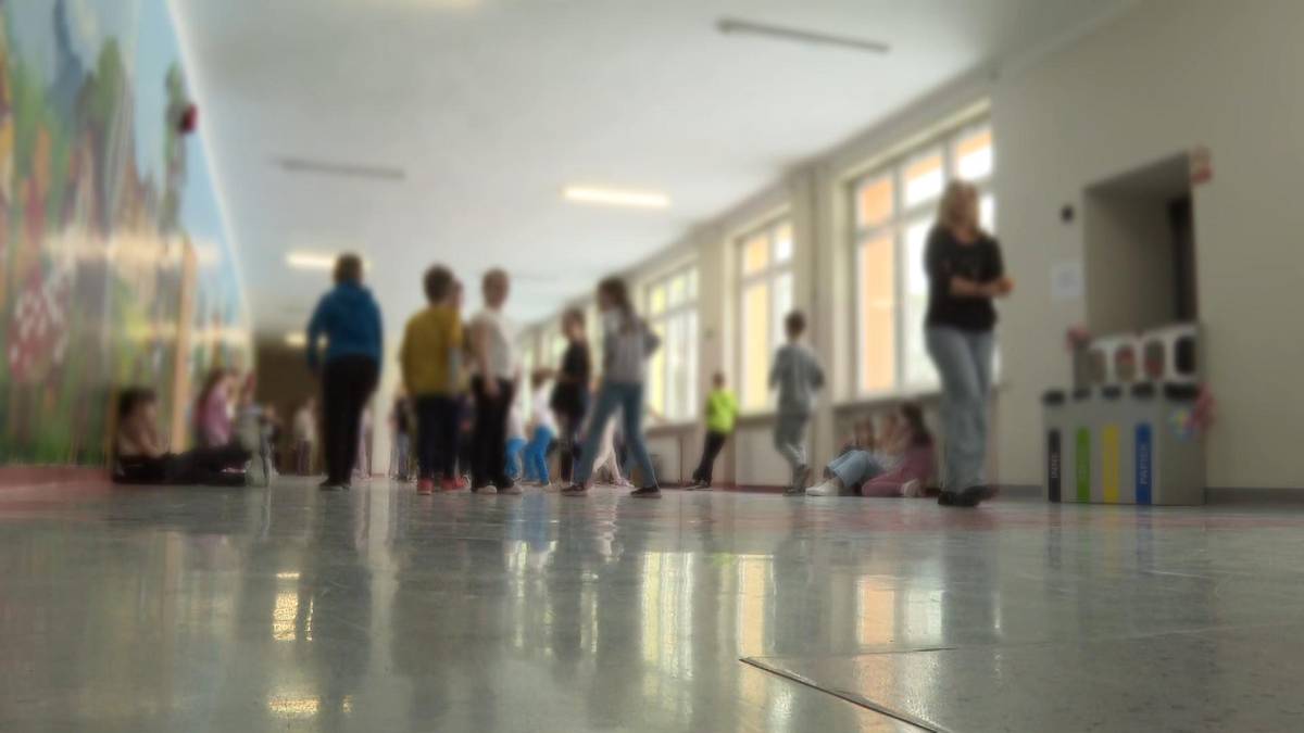 Grupa dzieci i dorosłych na korytarzu szkolnym, z oknami po prawej stronie i muralem na lewej ścianie.