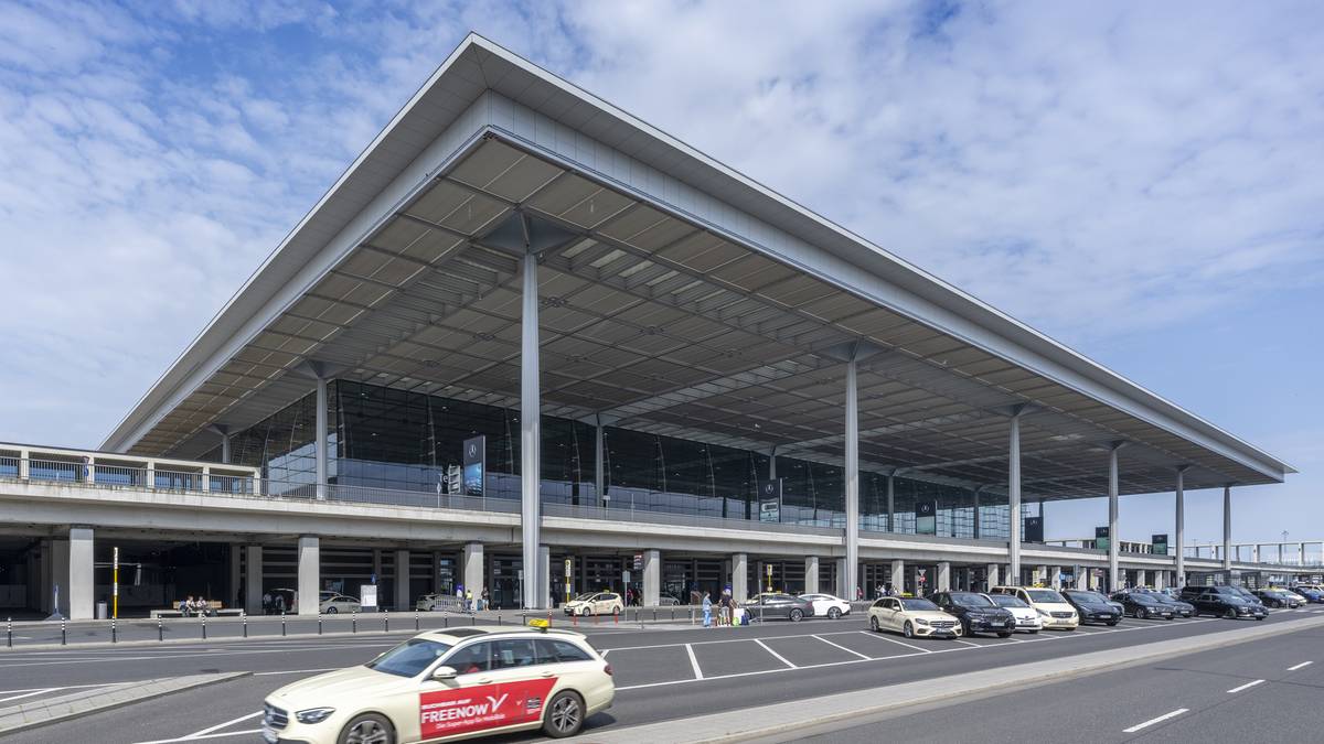 Widok na nowoczesny budynek terminala lotniska Berlin-Brandenburg z charakterystycznym, szerokim zadaszeniem. Przed budynkiem znajduje się ulica z kilkoma samochodami, w tym biała taks&oacute;wka z czerwonym logo.