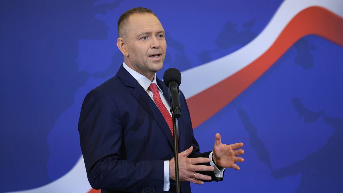 Mężczyzna w garniturze i krawacie przemawia przy mikrofonie na tle z biało-czerwonymi pasami.