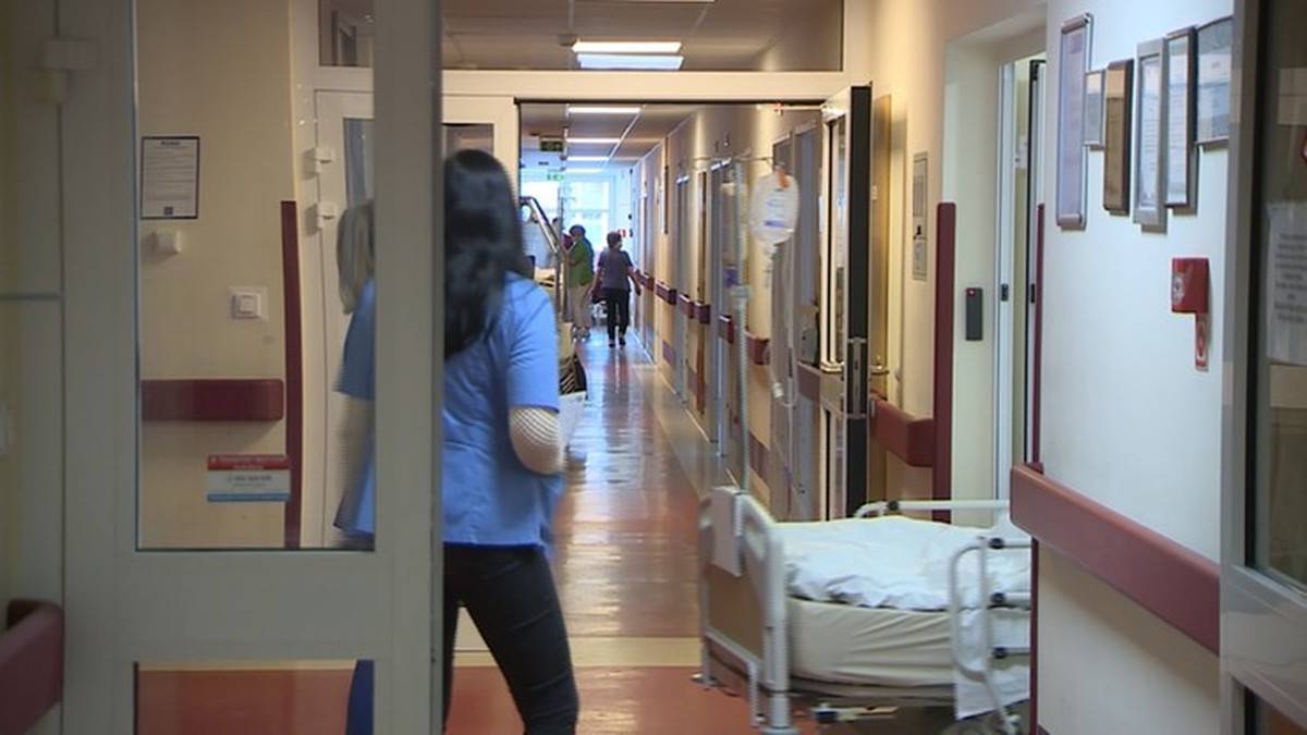 Korytarz szpitalny z ł&oacute;żkiem pacjenta i personelu medycznego w tle.