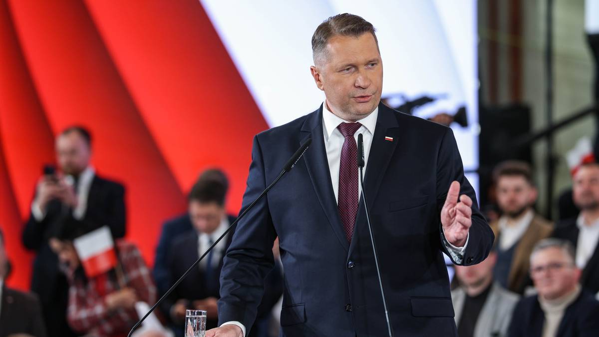 Mężczyzna w garniturze przemawia do mikrofonu na tle czerwono-białej scenografii.