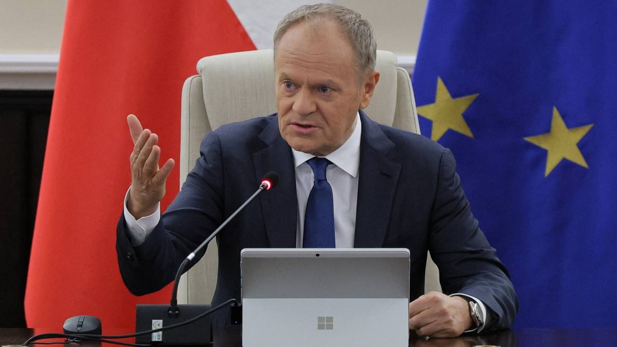Premier Donald Tusk przemawia, siedząc za biurkiem z laptopem. W tle widoczne flagi Polski i Unii Europejskiej.