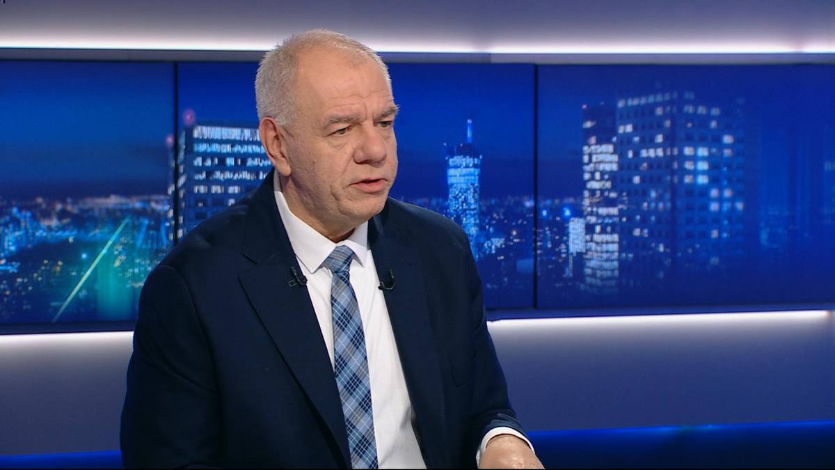 Mężczyzna w średnim wieku w garniturze i krawacie siedzi przed ekranem z widokiem nocnego miasta.