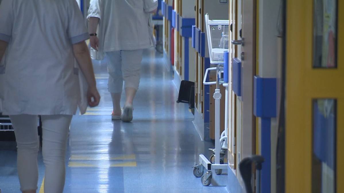 Sylwetki dw&oacute;ch os&oacute;b w białych fartuchach idących długim korytarzem szpitalnym.