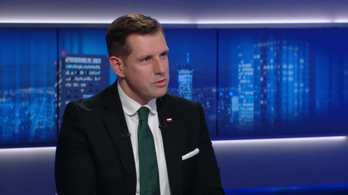 Mężczyzna w garniturze i krawacie siedzi i m&oacute;wi, w tle widać nocną panoramę miasta.