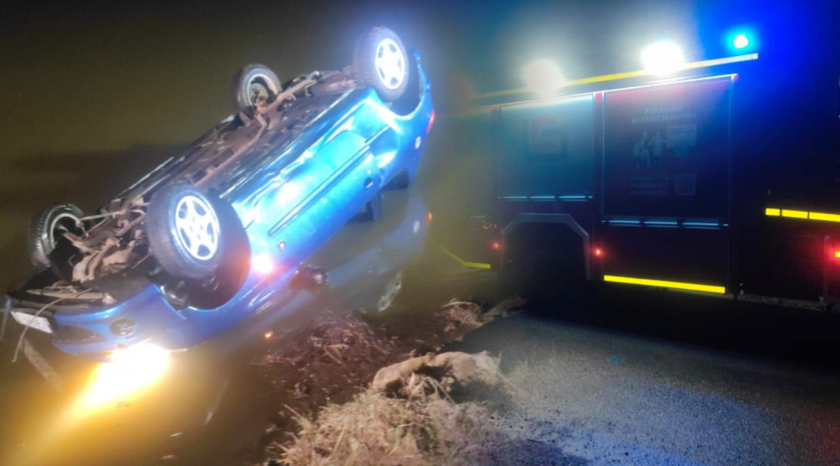 Przewr&oacute;cony niebieski samoch&oacute;d osobowy na poboczu drogi nocą, obok widoczny w&oacute;z strażacki z włączonymi sygnałami świetlnymi.