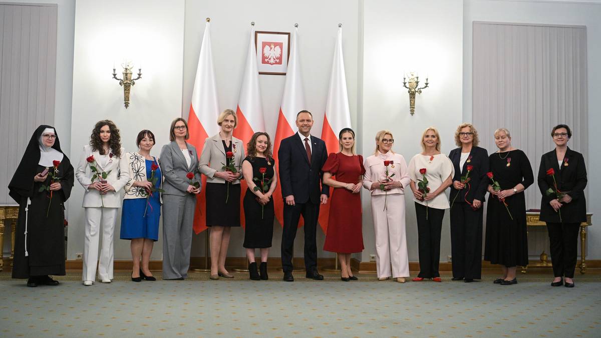 Grupa os&oacute;b, w tym mężczyzna w garniturze i kobiety w formalnych strojach, stoi w rzędzie. Niekt&oacute;re osoby trzymają r&oacute;że. W tle wiszą flagi i obraz.