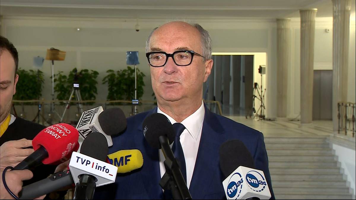 Mężczyzna w garniturze i okularach przemawia do mikrofon&oacute;w.