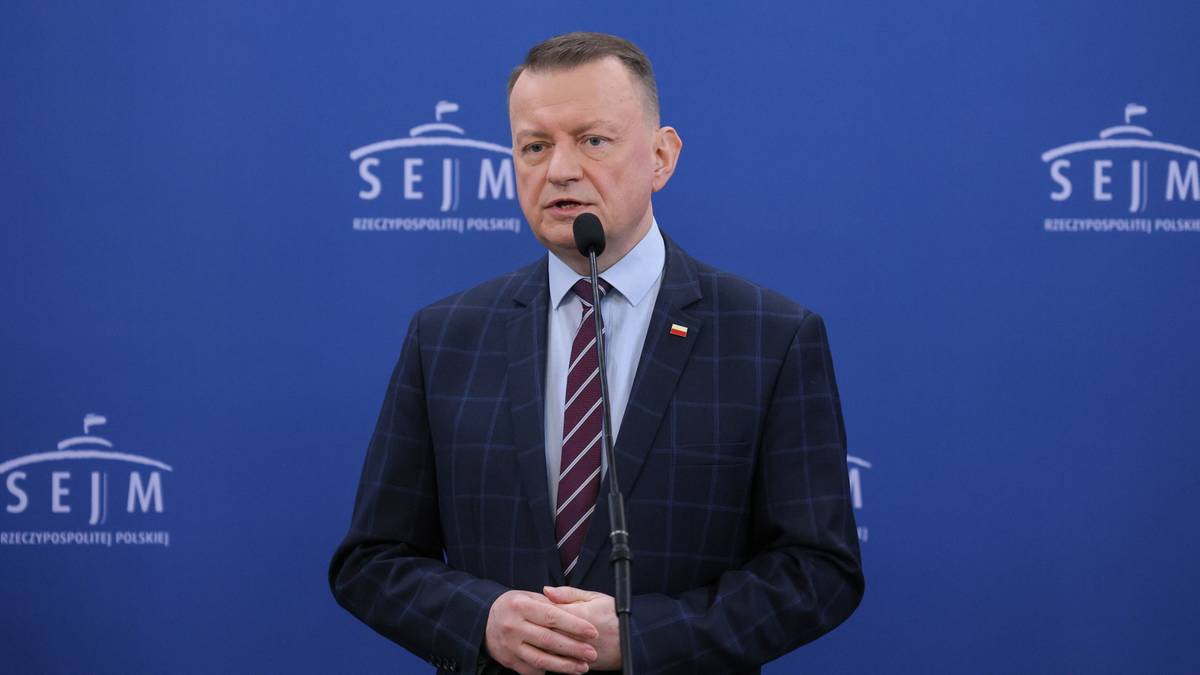 Polityk w garniturze stoi przy mikrofonie na tle z logo Sejmu RP.