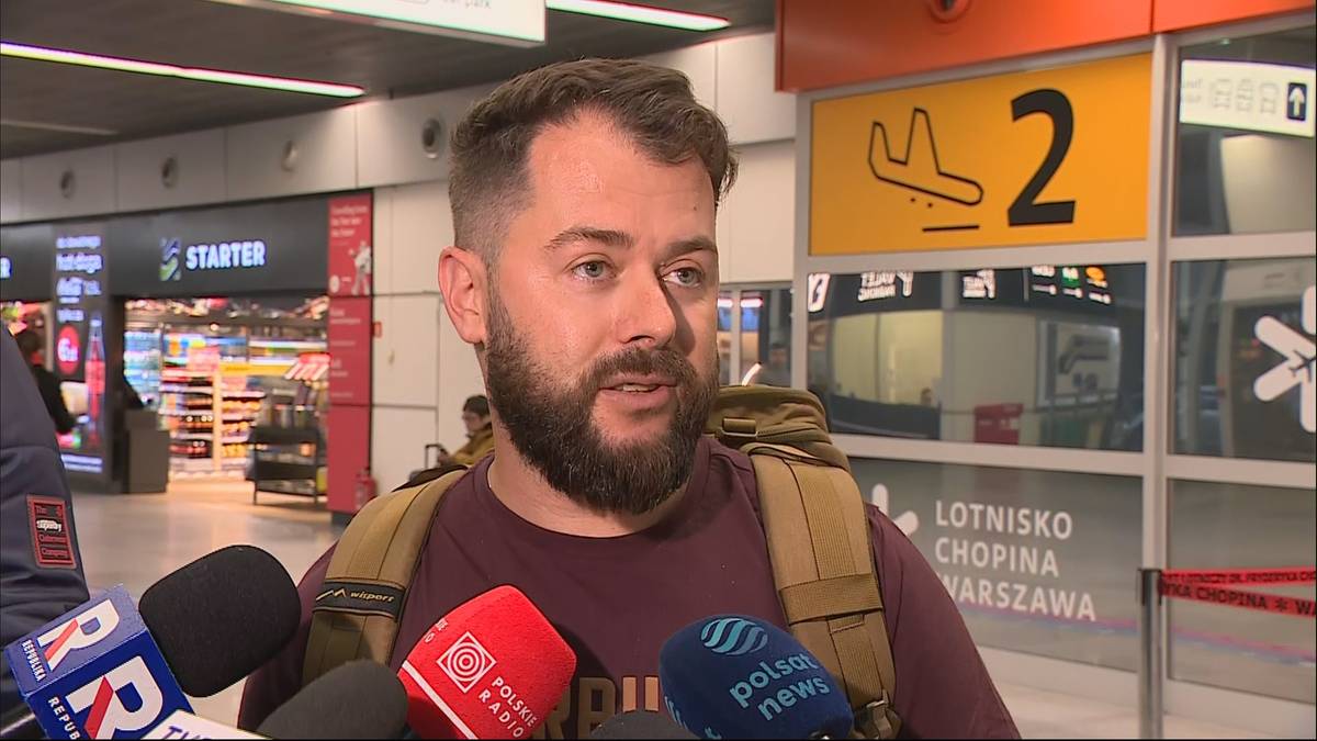 Mężczyzna z brodą, ubrany w ciemną koszulkę i plecak, odpowiada na pytania dziennikarzy przed mikrofonami.