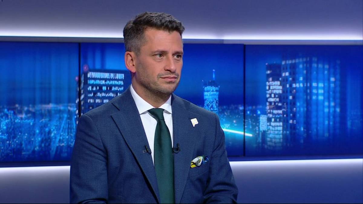 Mężczyzna w garniturze i krawacie siedzi przed ekranami z widokiem nocnego miasta.