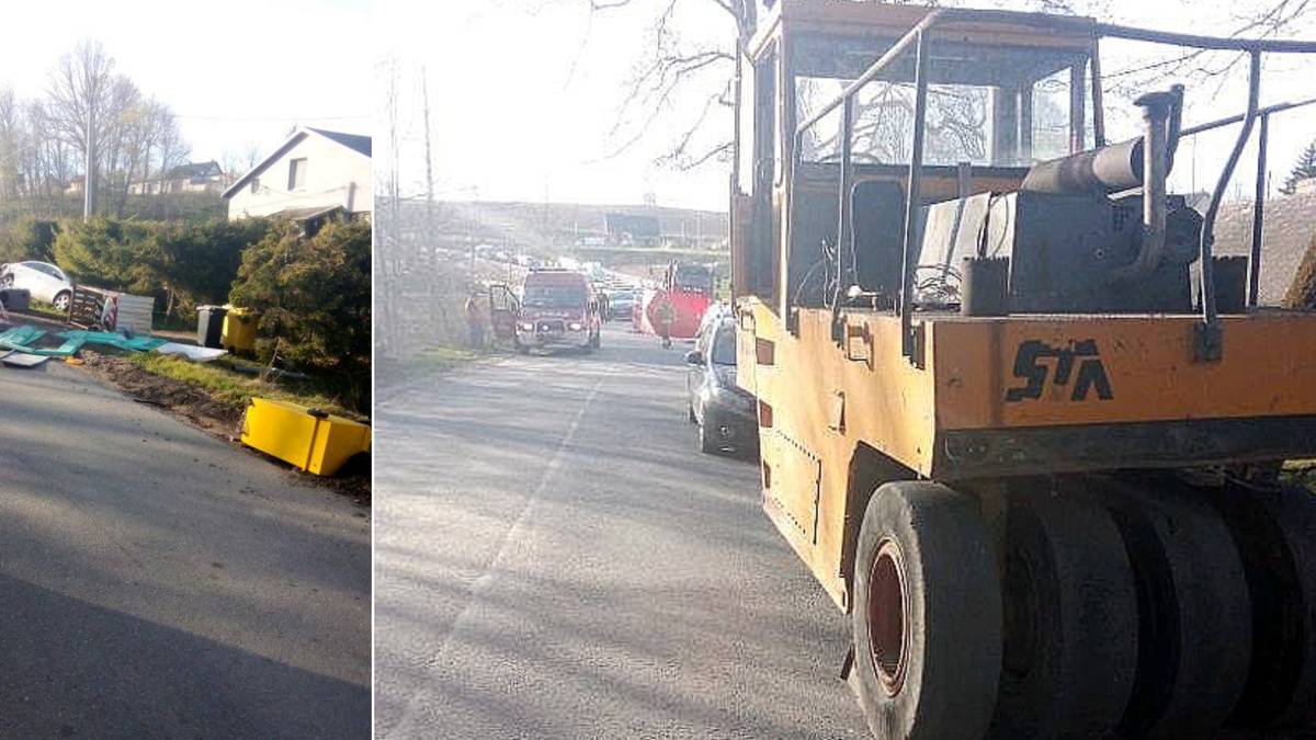 Zdjęcie przedstawia walec drogowy stojący na poboczu drogi. W tle widoczne są inne pojazdy oraz zabudowania.