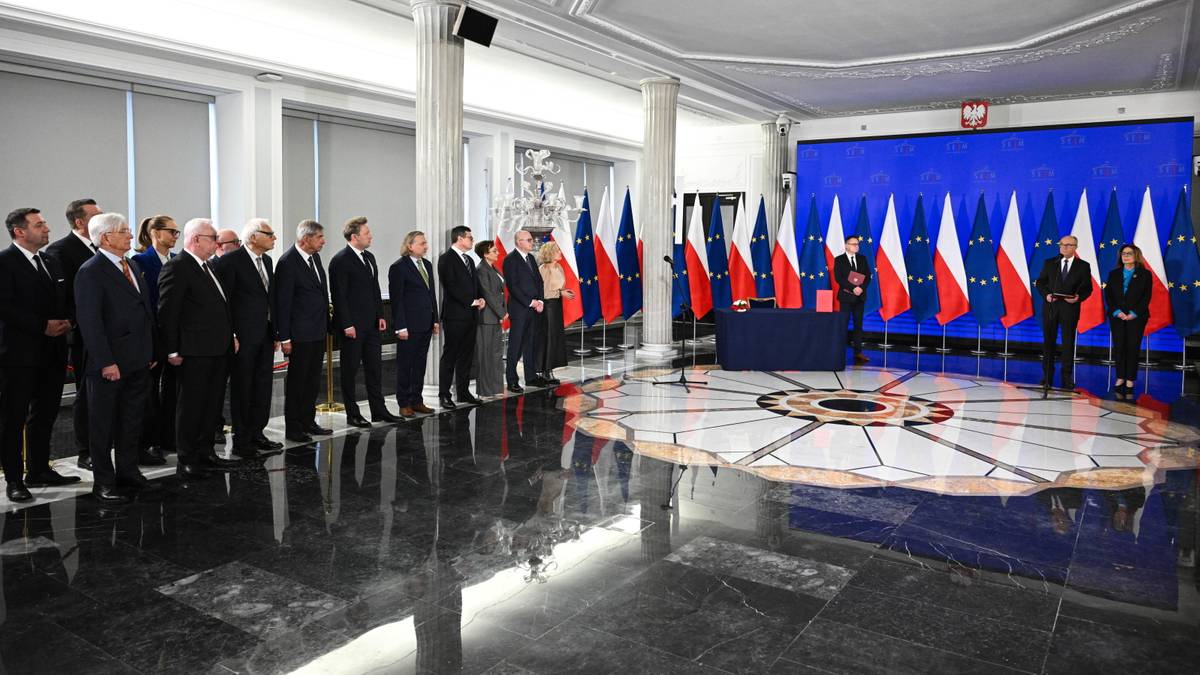 Grupa mężczyzn w garniturach stoi w szeregu przed flagami Polski i Unii Europejskiej, przed nimi znajduje się podium i m&oacute;wnica.