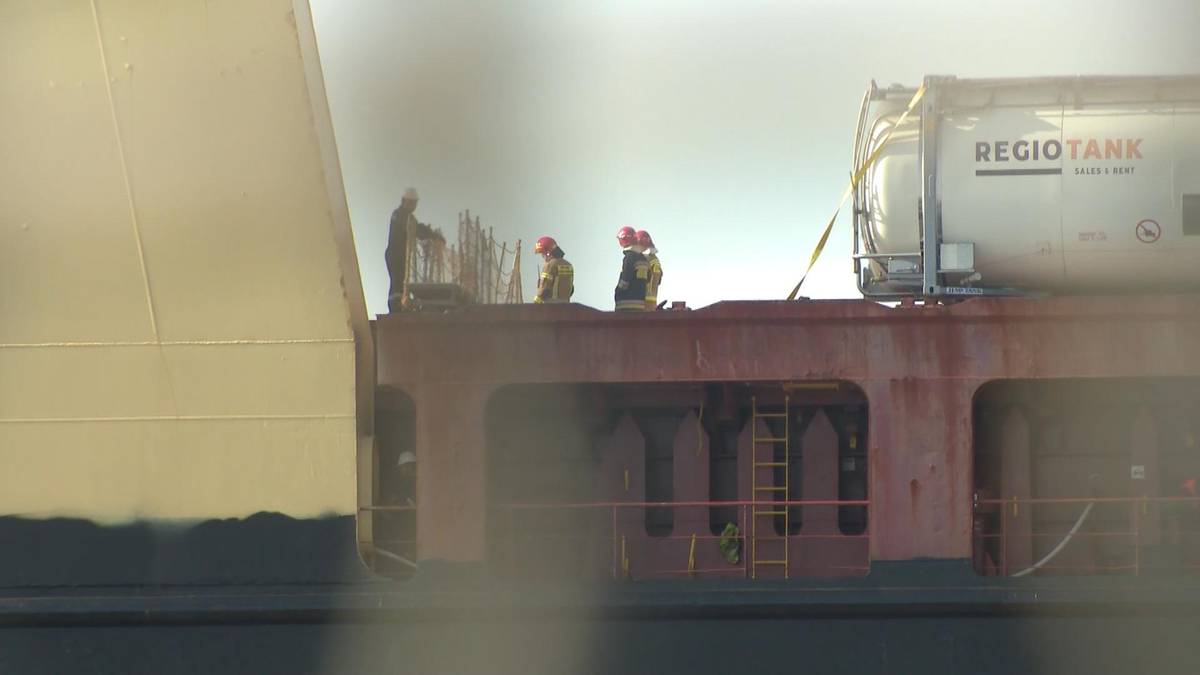 Widok fragmentu statku towarowego z kilkoma osobami w kamizelkach ratunkowych stojącymi na pokładzie, na tle jasnego nieba.