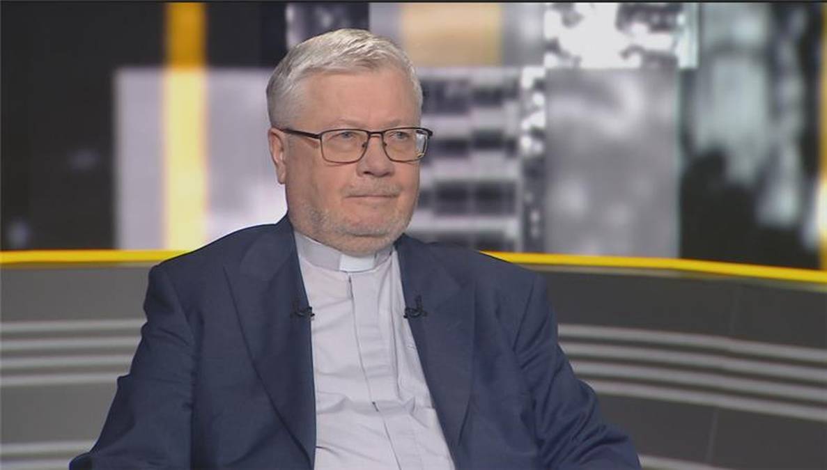 Ksiądz Alfred Wierzbicki w ciemnym garniturze i białej koszuli z duchownym kołnierzykiem, siedzi w studiu telewizyjnym.