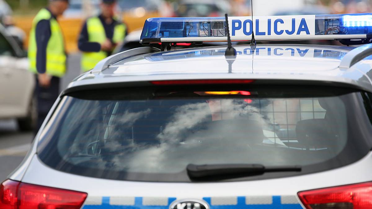 Zagadkowy zgon w Bystrzycy Kłodzkiej. Dwie osoby aresztowane. 3 Widok z tyłu na radiowóz policyjny z zapalonymi niebieskimi światłami i napisem "POLICJA". W tle widać dwóch policjantów w kamizelkach odblaskowych.