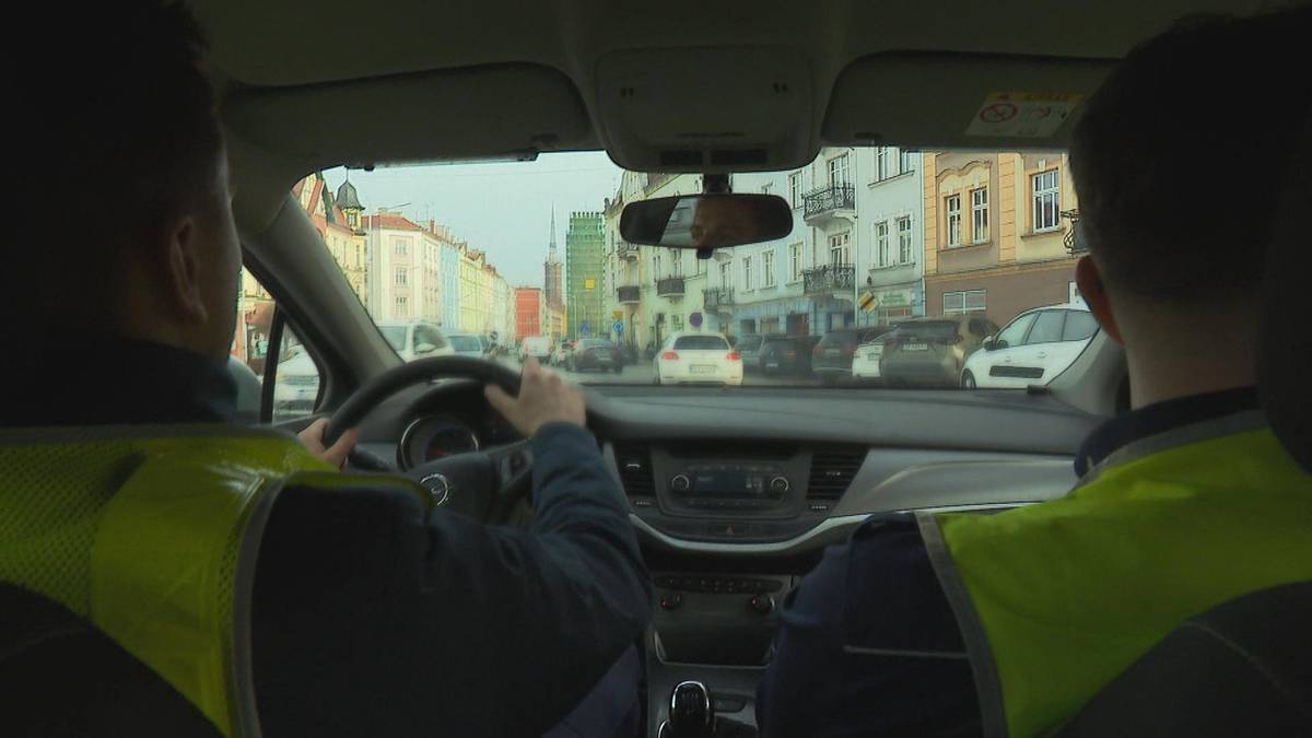 Dw&oacute;ch policjant&oacute;w w kamizelkach odblaskowych siedzi w samochodzie policyjnym. Jeden z nich prowadzi, patrząc na drogę przed sobą.