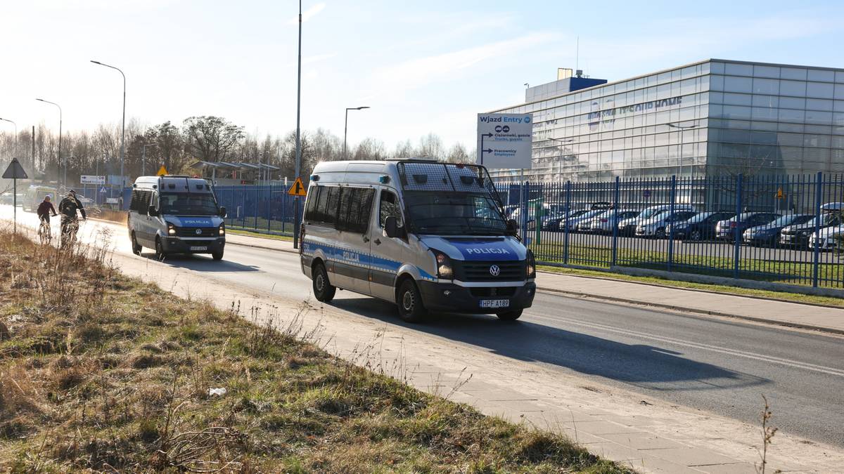 Samochody policyjne jadące drogą obok nowoczesnego budynku fabryki.
