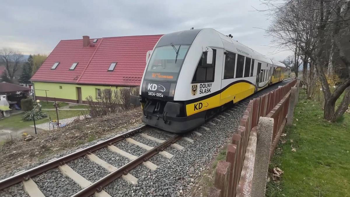 Nowoczesny pociąg Kolei Dolnośląskich z białym i ż&oacute;łtym malowaniem jedzie po torach obok zabudowań mieszkalnych.