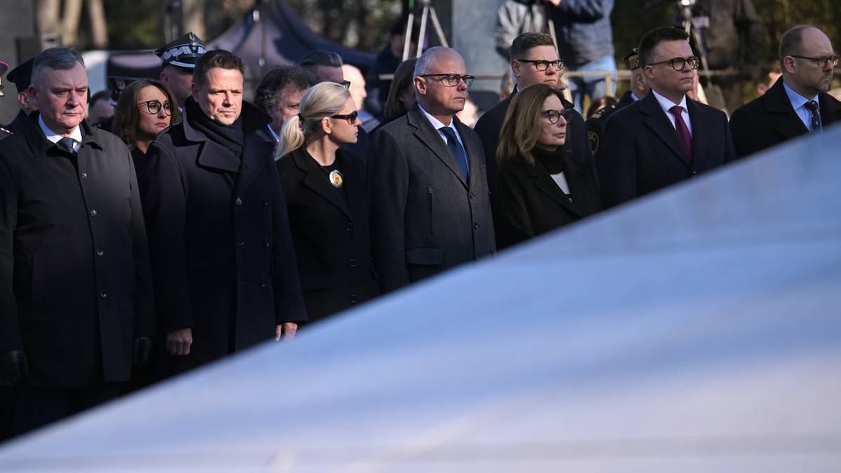 Grupa os&oacute;b w odświętnych strojach, stojących w uroczystej atmosferze na cmentarzu, prawdopodobnie podczas obchod&oacute;w rocznicowych.