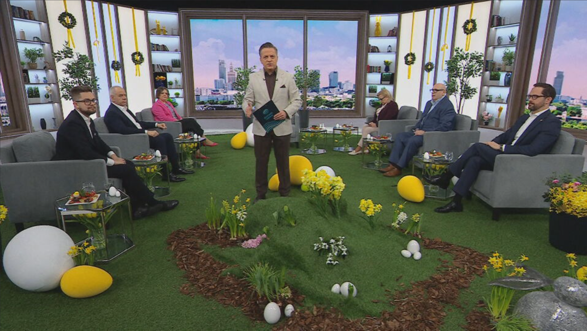 Prezenter stoi na środku scenografii z wiosennymi dekoracjami, otoczony przez siedzących gości.