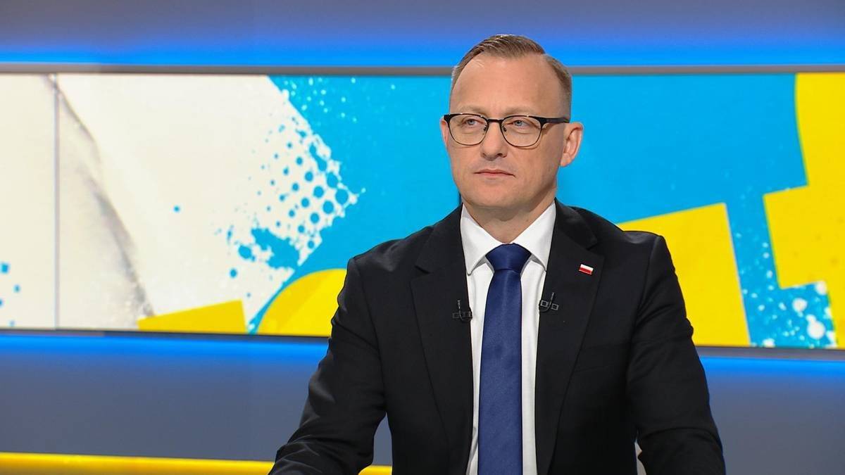 Mężczyzna w okularach, czarnym garniturze i granatowym krawacie siedzi i patrzy w bok. W tle znajduje się jasny ekran z abstrakcyjnym wzorem w kolorach niebieskim, białym i ż&oacute;łtym.