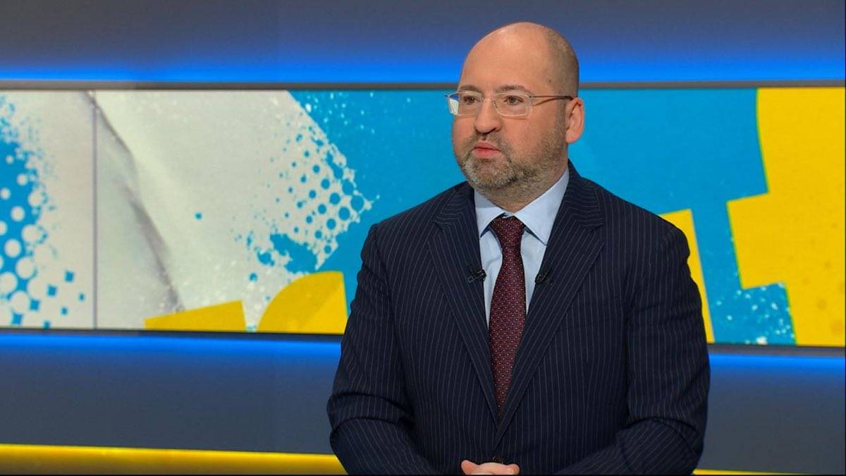 Mężczyzna w granatowym pasiastym garniturze, białej koszuli i bordowym krawacie, z okularami na nosie i br&oacute;dką, siedzi na tle studia telewizyjnego z niebiesko-ż&oacute;łtymi elementami graficznymi.