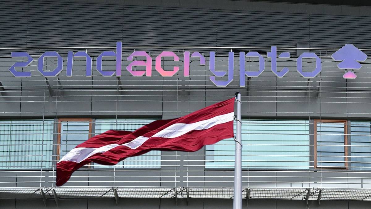 Flaga Łotwy powiewa na wietrze przed szarym budynkiem z neonowym, wielokolorowym napisem "zondacrypto".