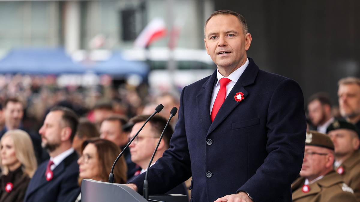 Mężczyzna w eleganckim stroju przemawia za m&oacute;wnicą, w tle widoczni ludzie.