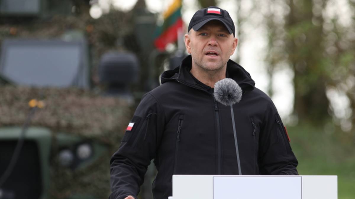 Mężczyzna w czapce z polską flagą przemawia przy m&oacute;wnicy z polską flagą.