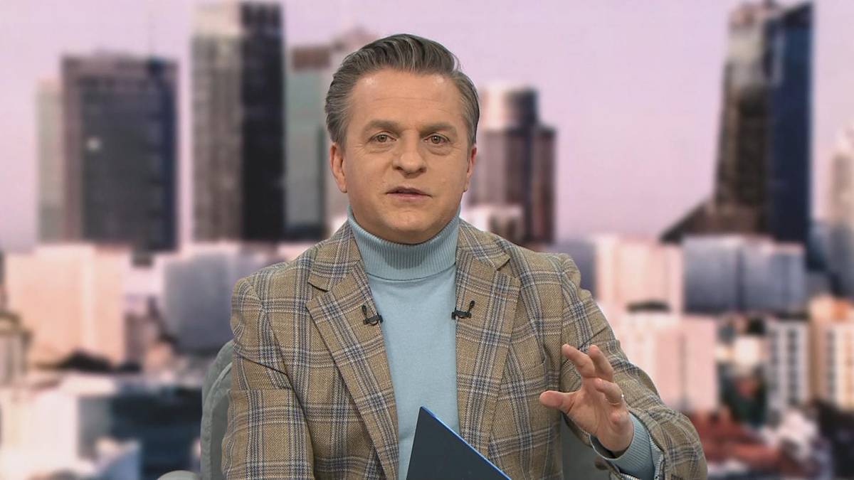 Mężczyzna w średnim wieku w eleganckim garniturze i golfie, siedzący w studiu telewizyjnym i gestykulujący podczas rozmowy.