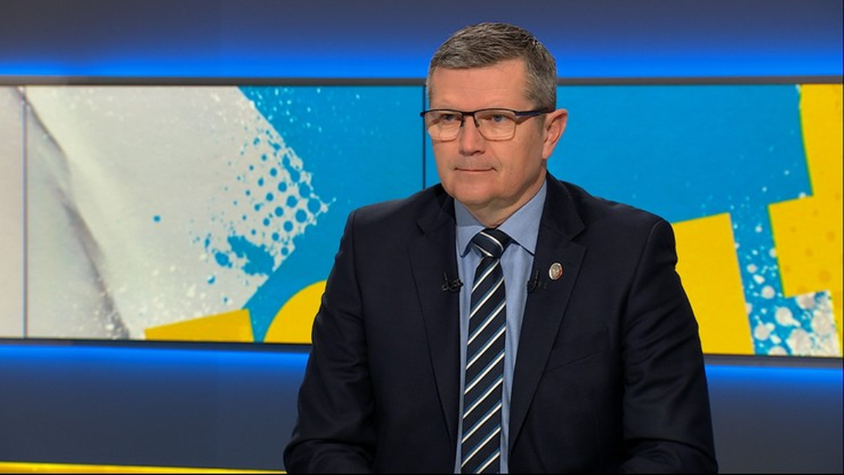 Mężczyzna w ciemnym garniturze, białej koszuli i pasiastym krawacie, noszący okulary, siedzi w studiu telewizyjnym. Na ekranie w tle widoczny jest abstrakcyjny wz&oacute;r w kolorach niebieskim i ż&oacute;łtym.