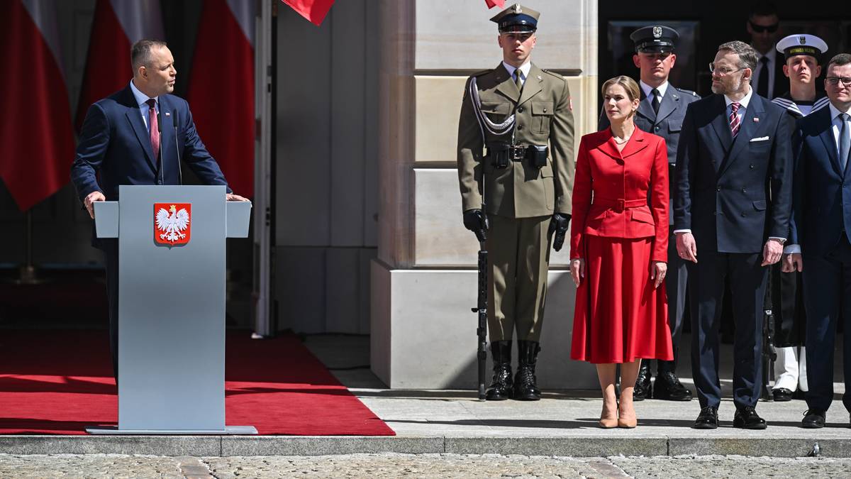 Mężczyzna przemawia za m&oacute;wnicą z godłem Polski, obok niego stoi kobieta w czerwonym stroju oraz żołnierze i mężczyźni w garniturach, na tle flag Polski.